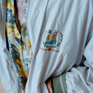 May include: A light gray jacket with a full zipper and a small embroidered emblem featuring two figures and the words "River Republic". The jacket is worn over a patterned button-down shirt. The sleeve cuff is green with pink stripes.