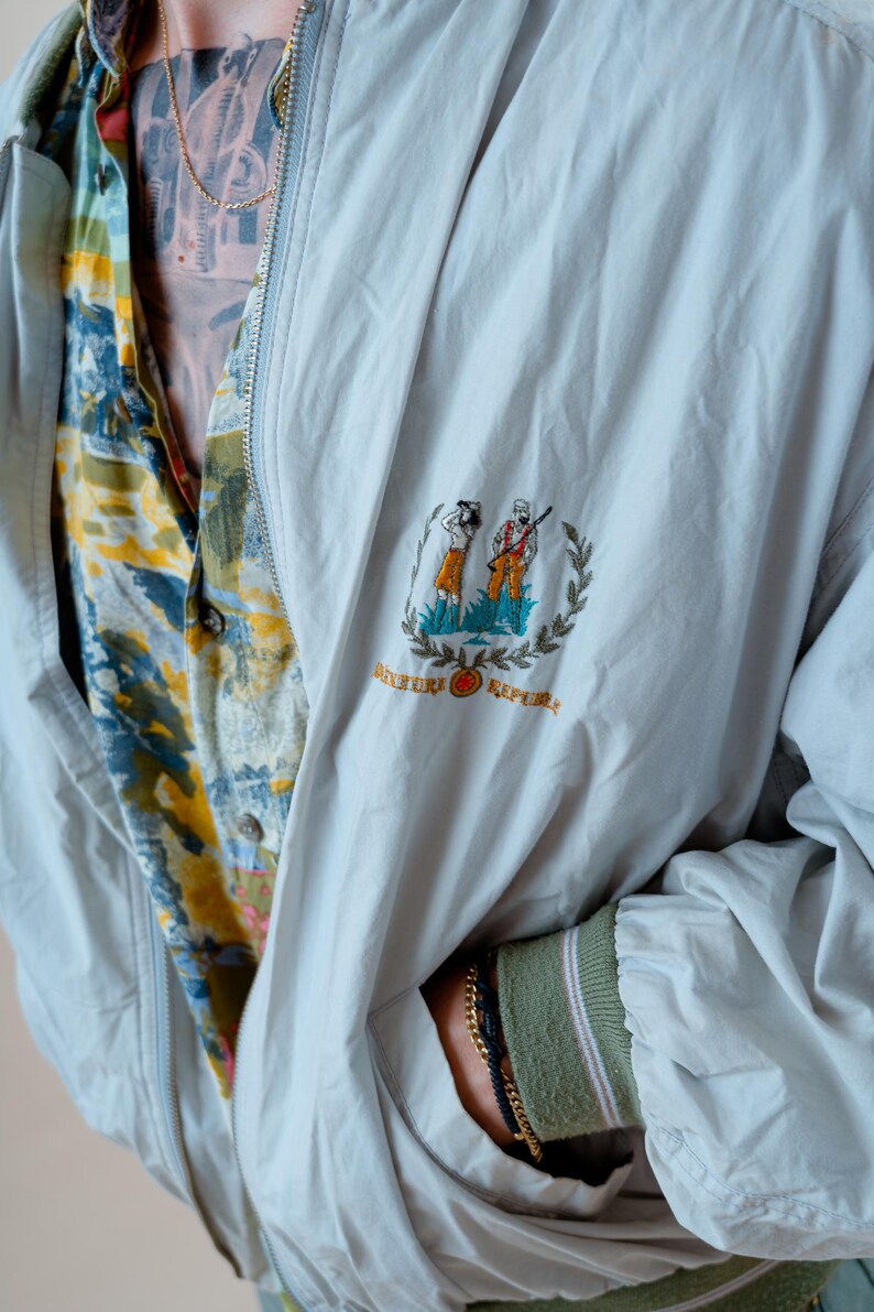 May include: A light gray jacket with a full zipper and a small embroidered emblem featuring two figures and the words "River Republic". The jacket is worn over a patterned button-down shirt. The sleeve cuff is green with pink stripes.