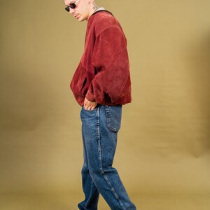 May include: A person wearing a rust-colored jacket, blue jeans, and white sneakers. The jacket has a relaxed fit. The jeans are a medium wash. The sneakers have black accents. The person is wearing sunglasses.