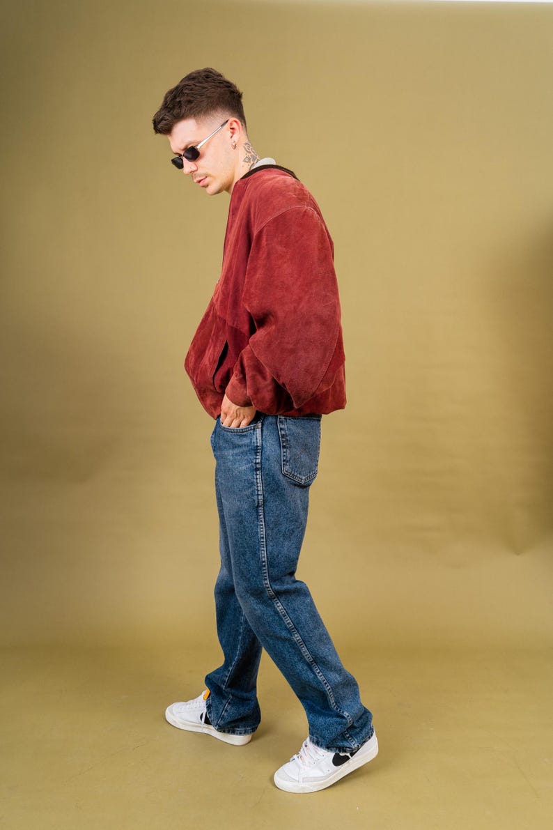 May include: A person wearing a rust-colored jacket, blue jeans, and white sneakers. The jacket has a relaxed fit. The jeans are a medium wash. The sneakers have black accents. The person is wearing sunglasses.