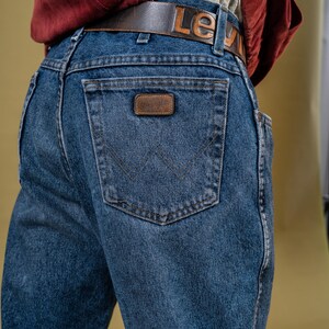 May include: Close-up of blue denim jeans with a brown leather belt. The belt has the word "Levi" embossed on it. The jeans feature a Wrangler patch on the back pocket.