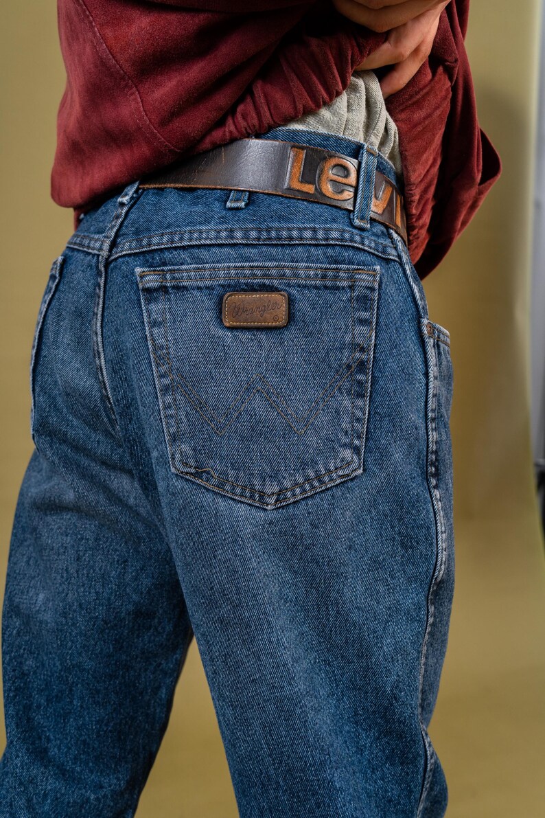 May include: Close-up of blue denim jeans with a brown leather belt. The belt has the word "Levi" embossed on it. The jeans feature a Wrangler patch on the back pocket.