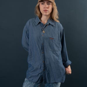 Könnte beinhalten: Eine Frau trägt ein blaues Jeanshemd mit einer braunen Baseballkappe und hellblaue Jeans. Das Hemd hat eine Tasche auf der linken Seite mit einem kleinen roten Logo.