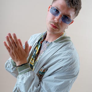 May include: A light blue bomber jacket with green trim, worn over a patterned shirt. The person is wearing blue-tinted sunglasses and a bracelet. The background is a solid beige color.