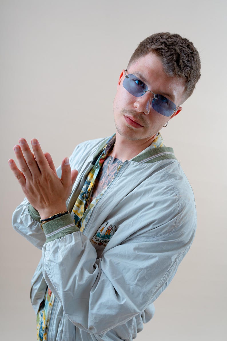 May include: A light blue bomber jacket with green trim, worn over a patterned shirt. The person is wearing blue-tinted sunglasses and a bracelet. The background is a solid beige color.