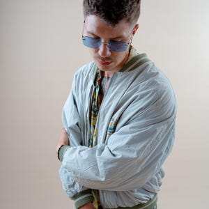 May include: A light blue bomber jacket with green and white trim, worn over a patterned shirt. The person is wearing blue-tinted sunglasses. The jacket has a small embroidered design on the chest. The person is also wearing blue jeans.