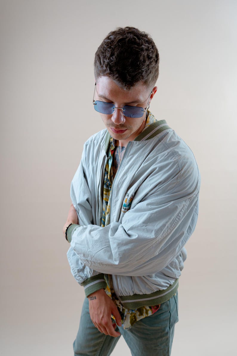 May include: A light blue bomber jacket with green and white trim, worn over a patterned shirt. The person is wearing blue-tinted sunglasses. The jacket has a small embroidered design on the chest. The person is also wearing blue jeans.