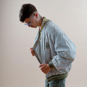 May include: A light blue bomber jacket with a green and white striped trim. The jacket has a small embroidered design on the chest. The person is wearing blue jeans and blue sunglasses.