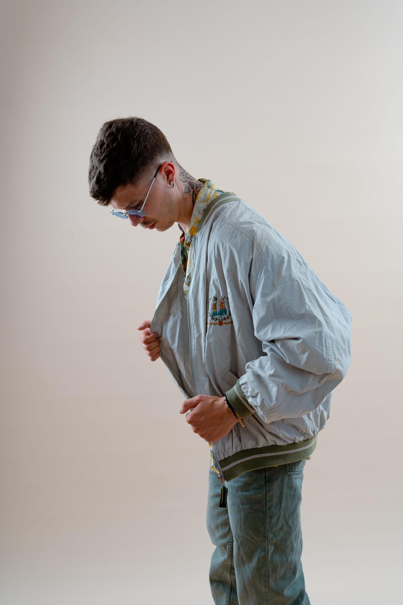 May include: A light blue bomber jacket with a green and white striped trim. The jacket has a small embroidered design on the chest. The person is wearing blue jeans and blue sunglasses.