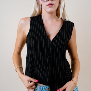 Könnte beinhalten: Eine schwarze Weste mit Nadelstreifen, V-Ausschnitt und Knopfverschluss. Die Weste wird mit einem Jeansrock und einer großen weißen Sonnenbrille mit dunklen Gläsern kombiniert. Das Model trägt mehrere Ringe und Armbänder.