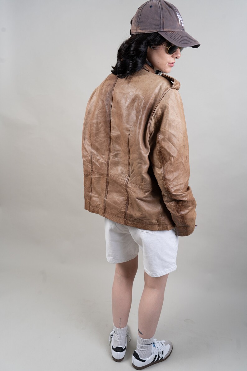 May include: A brown leather jacket with a white t-shirt underneath. The person is wearing white shorts and white trainers with black accents.