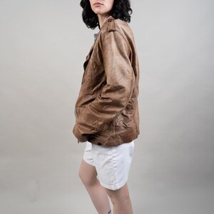 May include: A woman wearing a brown leather jacket, white shorts, and a black and white baseball cap with the letters "NY" on it. She is also wearing sunglasses and white trainers with black stripes.