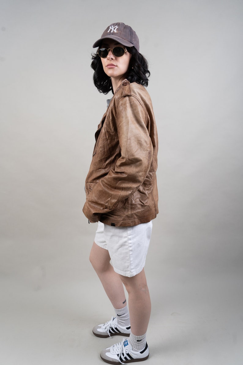 May include: A woman wearing a brown leather jacket, white shorts, and a black and white baseball cap with the letters "NY" on it. She is also wearing sunglasses and white trainers with black stripes.