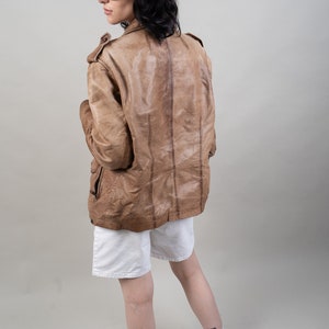 May include: A brown leather jacket with a zip fastening and a stand-up collar. The jacket is worn over a white t-shirt and white shorts. The person is wearing a grey baseball cap with a white logo and sunglasses.