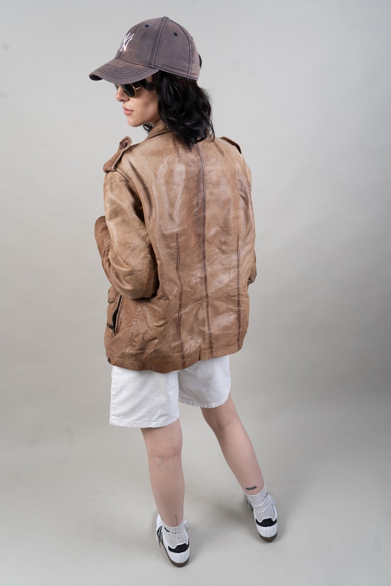 May include: A brown leather jacket with a zip fastening and a stand-up collar. The jacket is worn over a white t-shirt and white shorts. The person is wearing a grey baseball cap with a white logo and sunglasses.