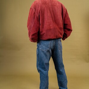 May include: A person wearing a red suede jacket with a dark collar and blue jeans. The person is also wearing white sneakers. The outfit is set against a solid olive-green background.