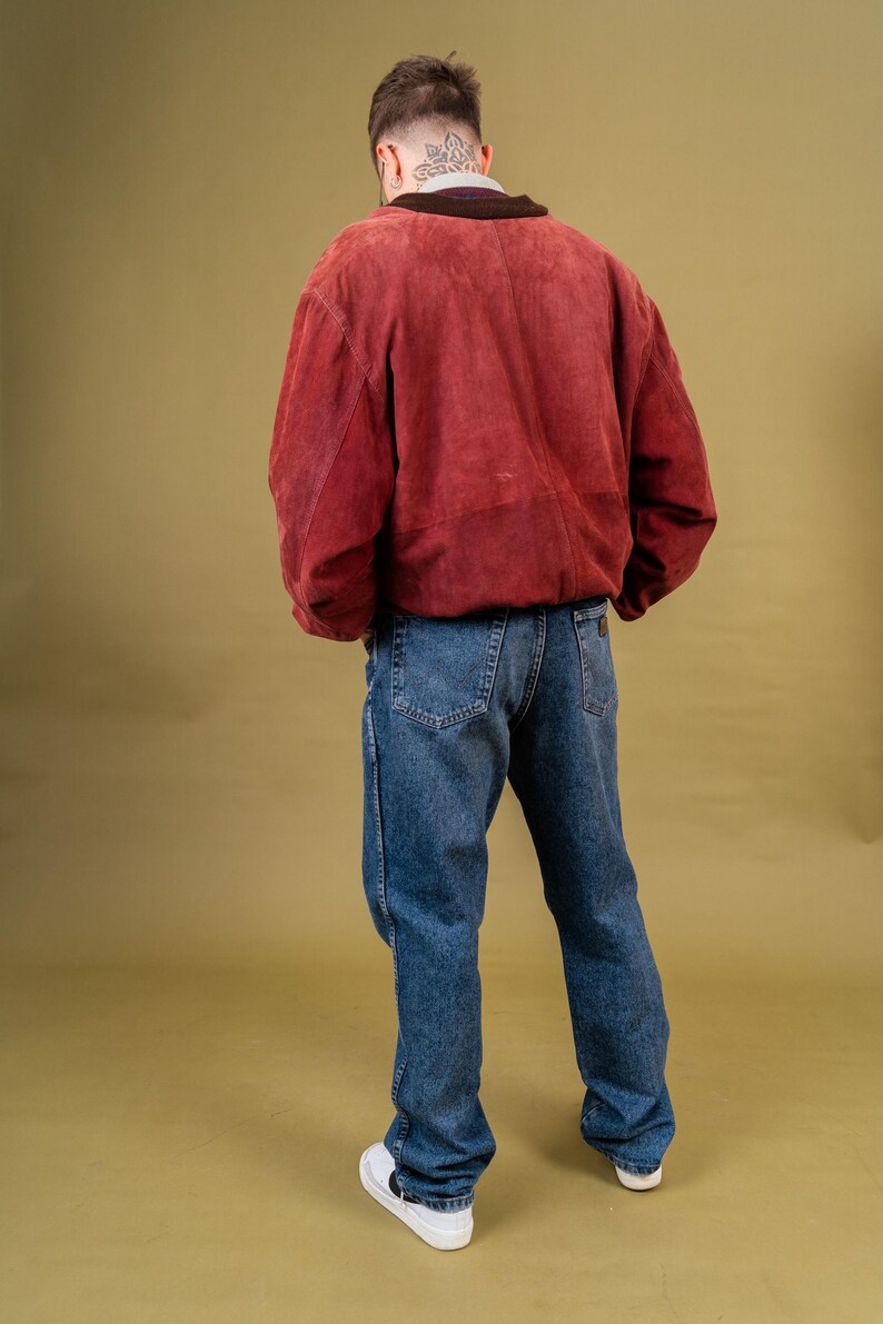 May include: A person wearing a red suede jacket with a dark collar and blue jeans. The person is also wearing white sneakers. The outfit is set against a solid olive-green background.