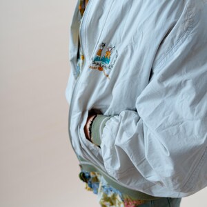 May include: A light blue bomber jacket with a colorful embroidered patch on the chest. The jacket has a zipper closure and a green collar and cuffs. The person is wearing a floral patterned shirt underneath.