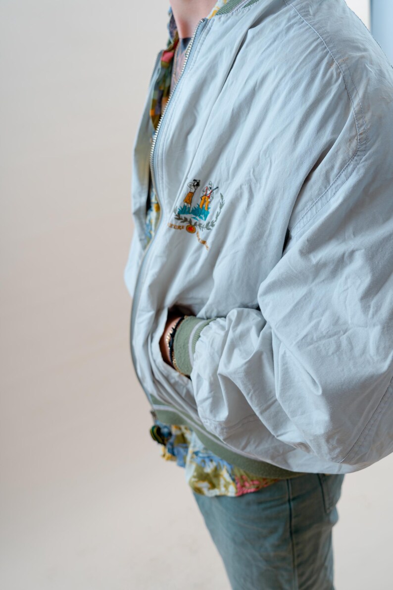 May include: A light blue bomber jacket with a colorful embroidered patch on the chest. The jacket has a zipper closure and a green collar and cuffs. The person is wearing a floral patterned shirt underneath.