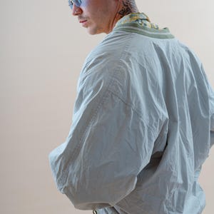 May include: A light blue bomber jacket with a green and white striped collar and cuffs. The jacket is worn over a patterned shirt. The model is wearing blue-tinted glasses.