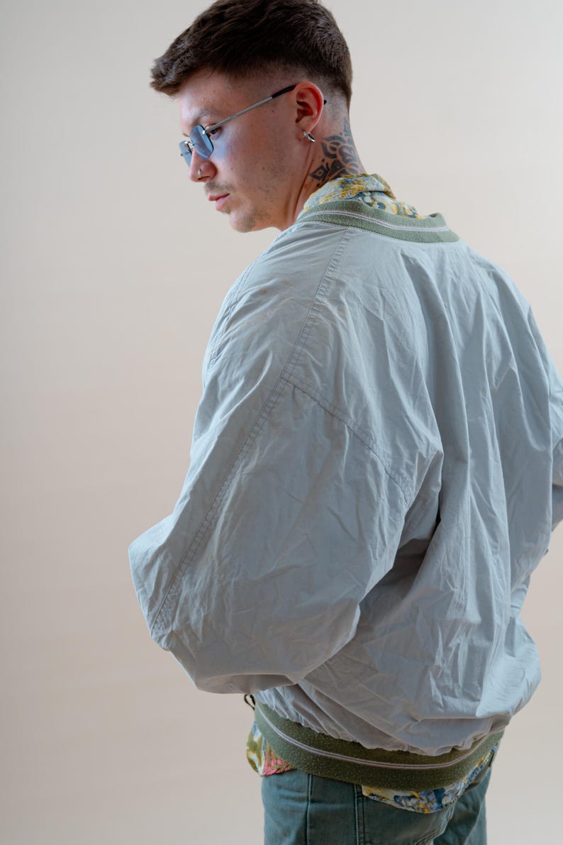 May include: A light blue bomber jacket with a green and white striped collar and cuffs. The jacket is worn over a patterned shirt. The model is wearing blue-tinted glasses.