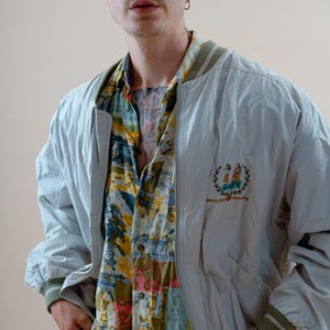 May include: A light blue bomber jacket with a green and white striped collar and cuffs. The jacket has a small embroidered emblem on the chest. Underneath, a colorful patterned shirt is visible.