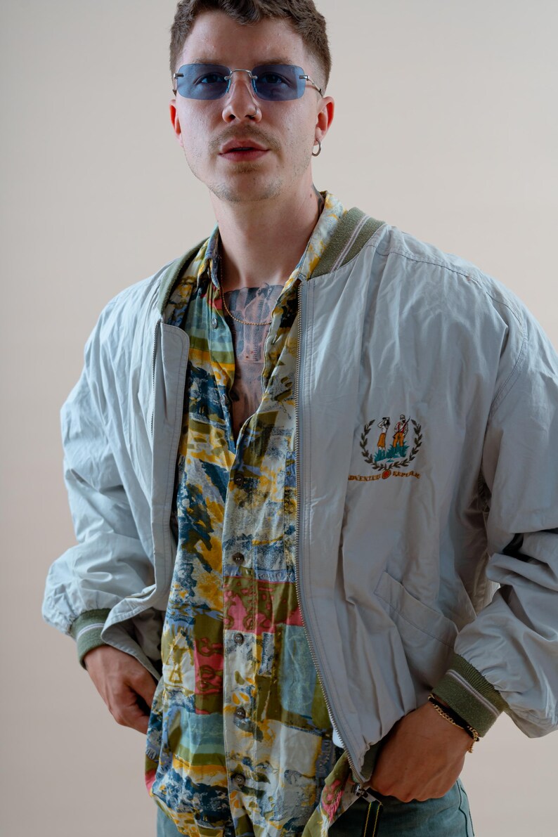 May include: A light blue bomber jacket with a green and white striped collar and cuffs. The jacket has a small embroidered emblem on the chest. Underneath, a colorful patterned shirt is visible.