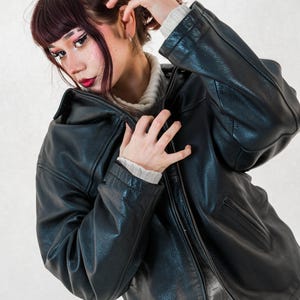 Könnte beinhalten: Schwarze Lederjacke mit Reißverschluss. Die Jacke hat ein klassisches Design mit Kragen und Taschen. Darunter wird ein hellfarbiger Pullover getragen. Die Jacke scheint in gutem Zustand zu sein und ist ein vielseitiges Kleidungsstück.