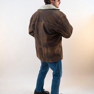 Op de afbeelding: Bruine su&egrave;de jas met een cr&egrave;mekleurige schapenvachtkraag. De jas wordt gecombineerd met een blauwe jeans en zwarte schoenen. Het model is van achteren te zien. Klassiek ontwerp.