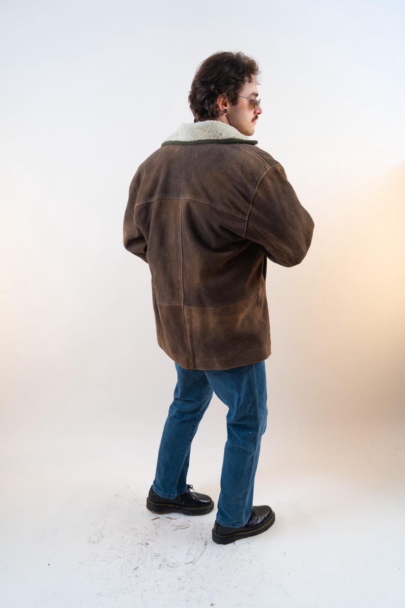Op de afbeelding: Bruine su&egrave;de jas met een cr&egrave;mekleurige schapenvachtkraag. De jas wordt gecombineerd met een blauwe jeans en zwarte schoenen. Het model is van achteren te zien. Klassiek ontwerp.