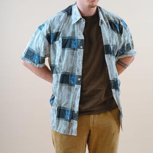Könnte beinhalten: Ein kurzärmeliges Hemd mit einem hellblauen, schwarzen und blauen Karomuster. Das Hemd ist offen und zeigt ein braunes T-Shirt darunter. Die Person trägt eine beigefarbene Cordhose.