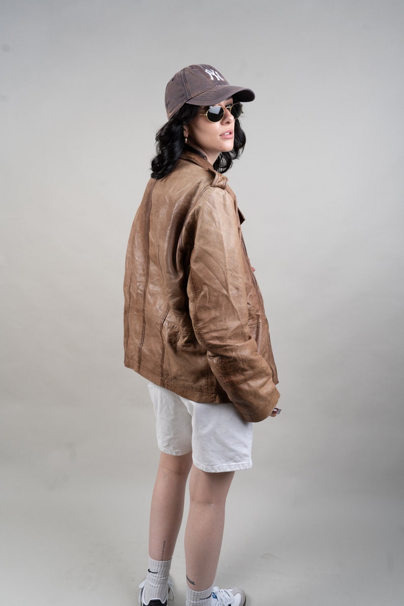May include: A woman wearing a brown leather jacket, white shorts, and a black baseball cap with the letters "NY" on it. She is also wearing sunglasses.