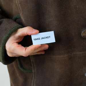 Op de afbeelding: Een bruine jas met houten knopen en groene biezen. De jas wordt in de hand van een persoon gehouden, met een klein wit label met de tekst "10452 JACKET". De jas lijkt gemaakt te zijn van een su&egrave;de-achtig materiaal.