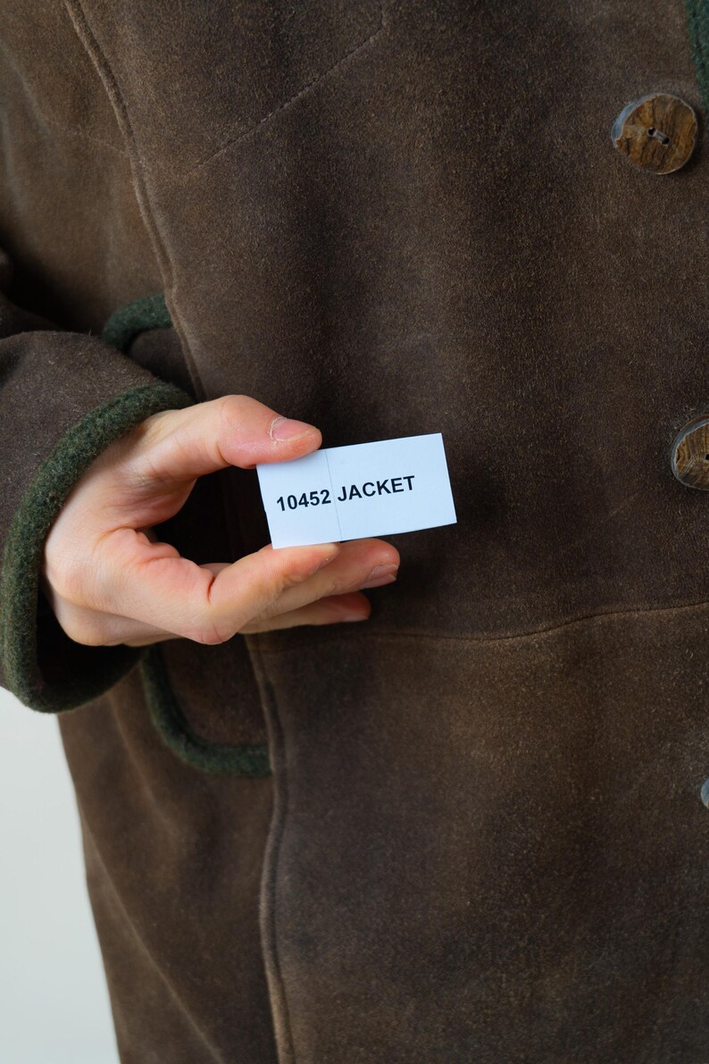 Op de afbeelding: Een bruine jas met houten knopen en groene biezen. De jas wordt in de hand van een persoon gehouden, met een klein wit label met de tekst "10452 JACKET". De jas lijkt gemaakt te zijn van een su&egrave;de-achtig materiaal.