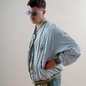 May include: A light blue bomber jacket with a green and white striped collar and cuffs. The jacket has a colorful embroidered patch on the chest. The person is wearing blue-tinted sunglasses and a patterned shirt.