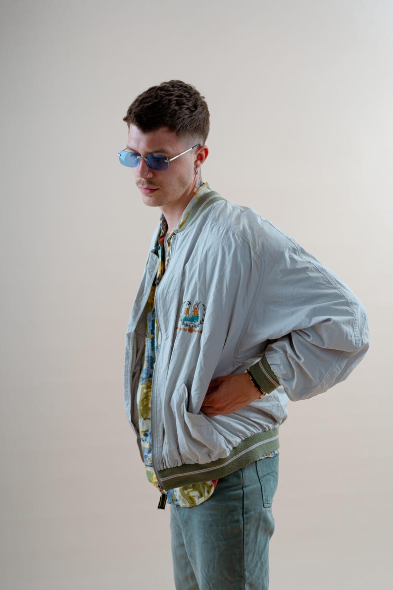 May include: A light blue bomber jacket with a green and white striped collar and cuffs. The jacket has a colorful embroidered patch on the chest. The person is wearing blue-tinted sunglasses and a patterned shirt.