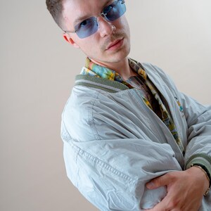 May include: A light gray bomber jacket with a green trim and a colorful patterned shirt underneath. The person is wearing blue-tinted rimless sunglasses. The jacket has a zipper closure and a small embroidered design on the chest.