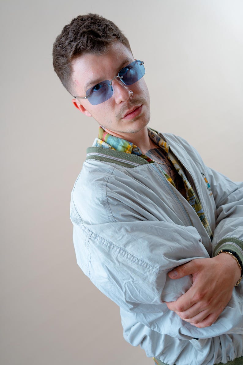 May include: A light gray bomber jacket with a green trim and a colorful patterned shirt underneath. The person is wearing blue-tinted rimless sunglasses. The jacket has a zipper closure and a small embroidered design on the chest.