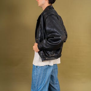 May include: A black leather bomber jacket with a ribbed hem and cuffs. The jacket is worn over a white sweater and blue striped pants. The person is wearing black sunglasses and white sneakers with green accents. The background is a solid olive green.