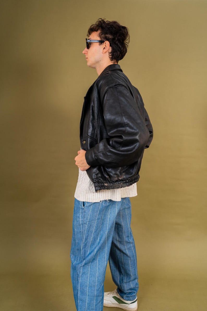 May include: A black leather bomber jacket with a ribbed hem and cuffs. The jacket is worn over a white sweater and blue striped pants. The person is wearing black sunglasses and white sneakers with green accents. The background is a solid olive green.