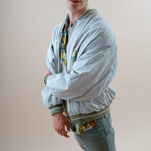 May include: A light blue bomber jacket with green trim, worn over a patterned shirt and paired with green pants. The person is wearing blue-tinted rimless sunglasses and black and white sneakers. The outfit is casual and stylish.