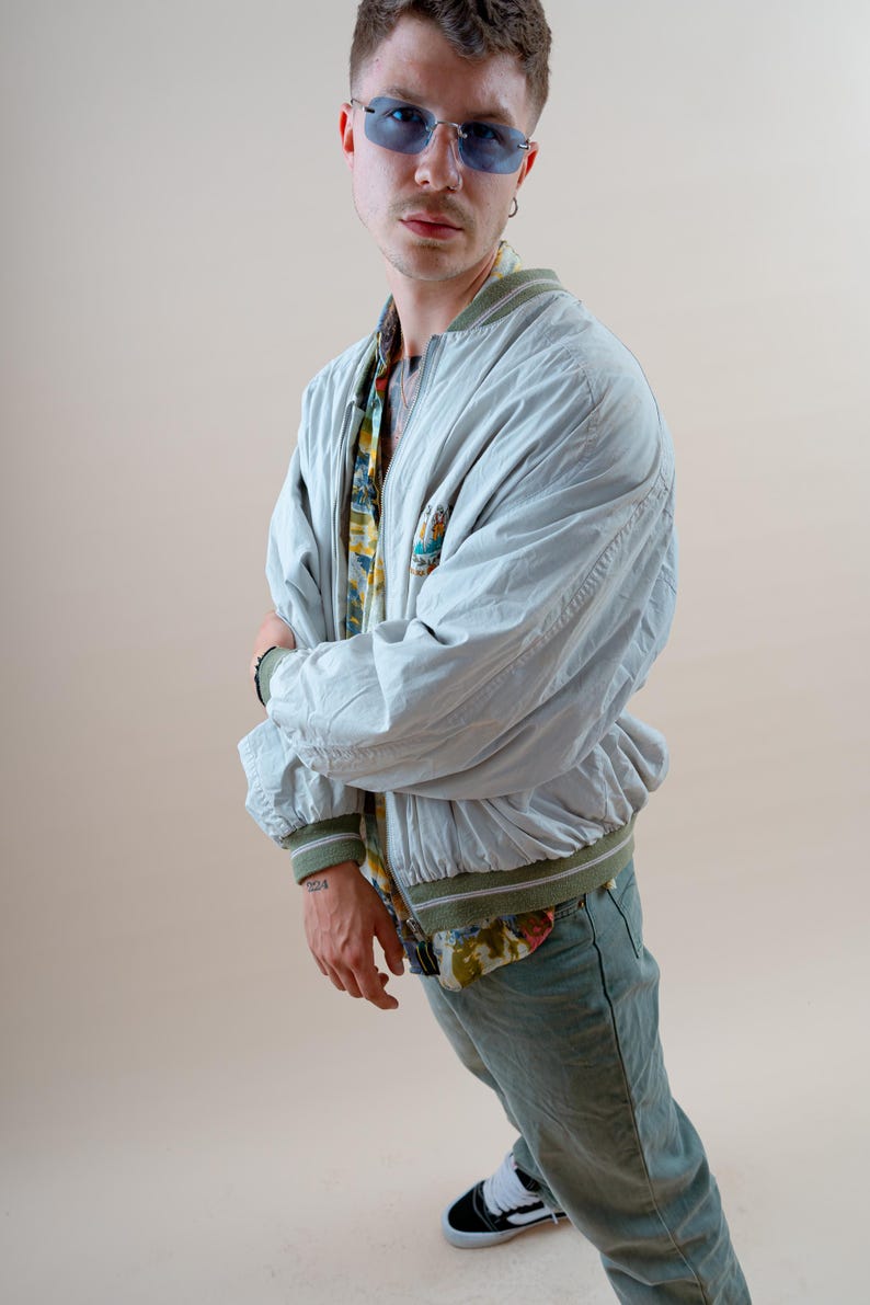 May include: A light blue bomber jacket with green trim, worn over a patterned shirt and paired with green pants. The person is wearing blue-tinted rimless sunglasses and black and white sneakers. The outfit is casual and stylish.