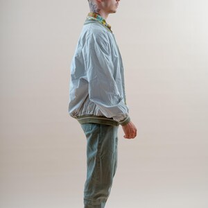 May include: A light gray bomber jacket with olive green ribbed trim, worn over a patterned collared shirt, paired with olive green trousers and black and white low-top sneakers. The outfit is completed with thin-framed glasses.