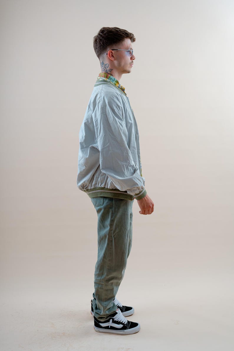 May include: A light gray bomber jacket with olive green ribbed trim, worn over a patterned collared shirt, paired with olive green trousers and black and white low-top sneakers. The outfit is completed with thin-framed glasses.