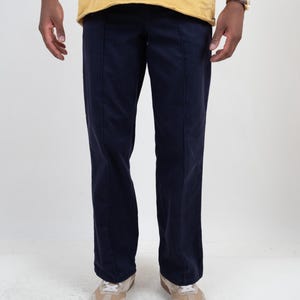 Könnte beinhalten: Eine dunkel marineblaue Hose mit geradem Schnitt und einer markanten vertikalen Bügelfalte in der Mitte jedes Hosenbeins. Die Hose hat eine schlichte, einfarbige Oberfläche und wird mit einem senfgelben T-Shirt kombiniert.