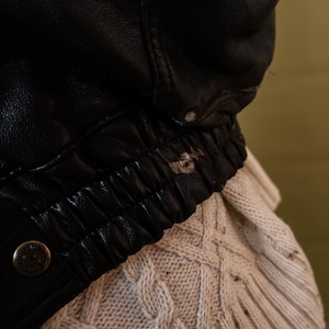 May include: Close-up of a black leather jacket with a ribbed elastic waistband and a button detail. The jacket is worn over a cream-colored cable-knit sweater, creating a contrast in textures and colors.