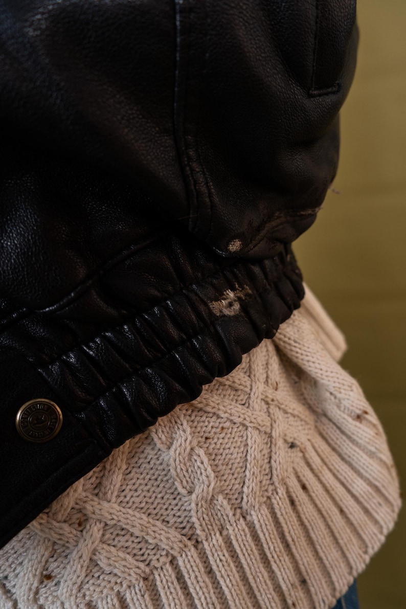 May include: Close-up of a black leather jacket with a ribbed elastic waistband and a button detail. The jacket is worn over a cream-colored cable-knit sweater, creating a contrast in textures and colors.