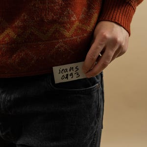 May include: A person wearing a red and yellow patterned sweater with a note that says "jeans 0493" tucked into the front pocket of their black jeans.