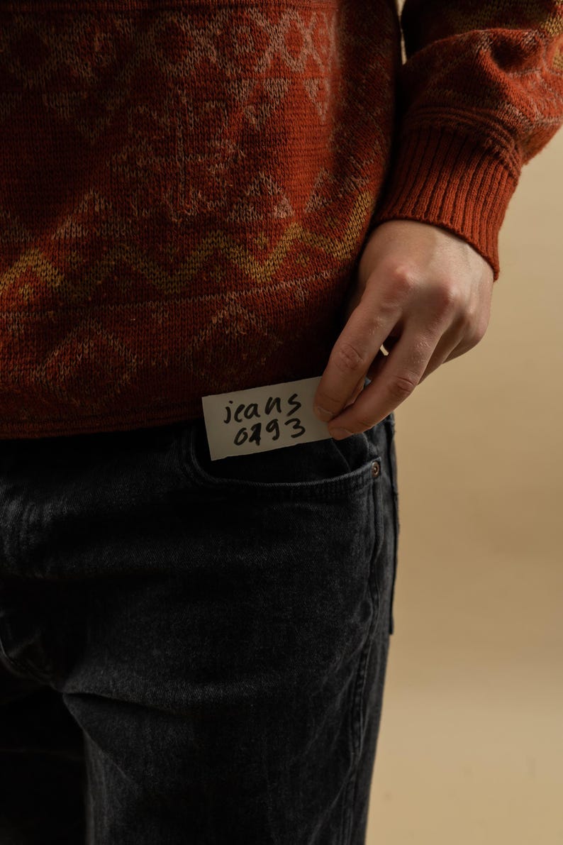 May include: A person wearing a red and yellow patterned sweater with a note that says "jeans 0493" tucked into the front pocket of their black jeans.
