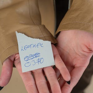 Pode incluir: Um close-up de uma jaqueta de couro marrom com uma nota escrita &agrave; m&atilde;o que diz "LEATHER" e tem os n&uacute;meros "036" e "0370" escritos nela.
